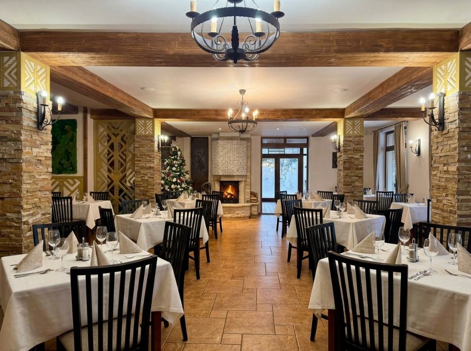 Sala de mese a restaurantului Domeniului Lidana, cu mese aranjate festiv, stâlpi placați cu piatră naturală și șemineu